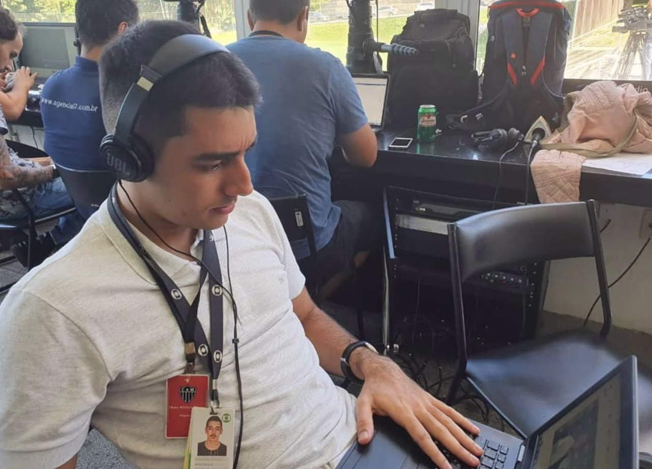 Sentado com um notebook no colo e um fone de ouvido, Frossard está com uma camisa branca e crachas pendurados no pescoço