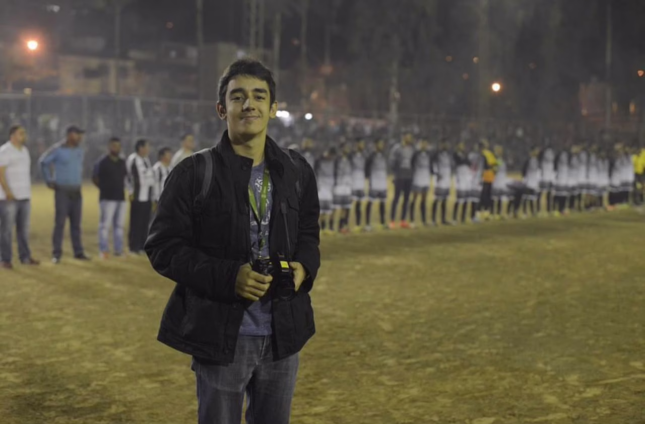 O jornalista está com uma blusa de frio preta e câmera na mão. Ao fundo, um campo de futebol com jogadores enfileirados