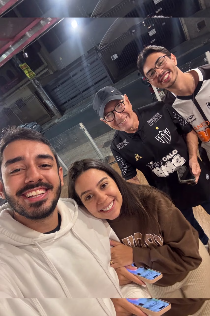 Um homem tira uma selfie com uma mulher segurando seu braço. Na foto, aparecem outros dois homens - um mais velho e outro jovem - com a camisa do Galo