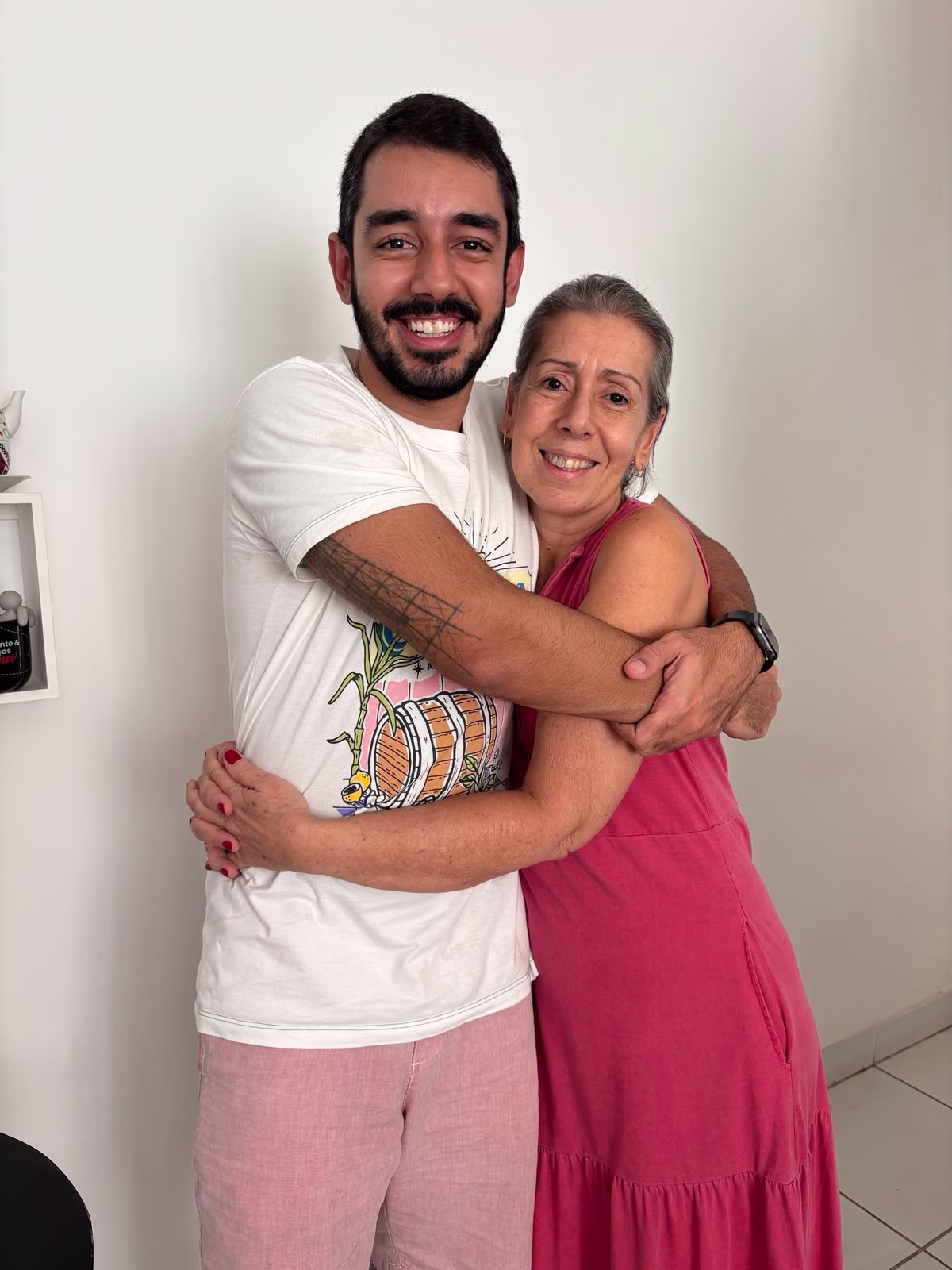 O homem de camisa branca aparece abraçado com uma mulher de cabelos curtos e grisalhos. Ela veste um vestido rosa
