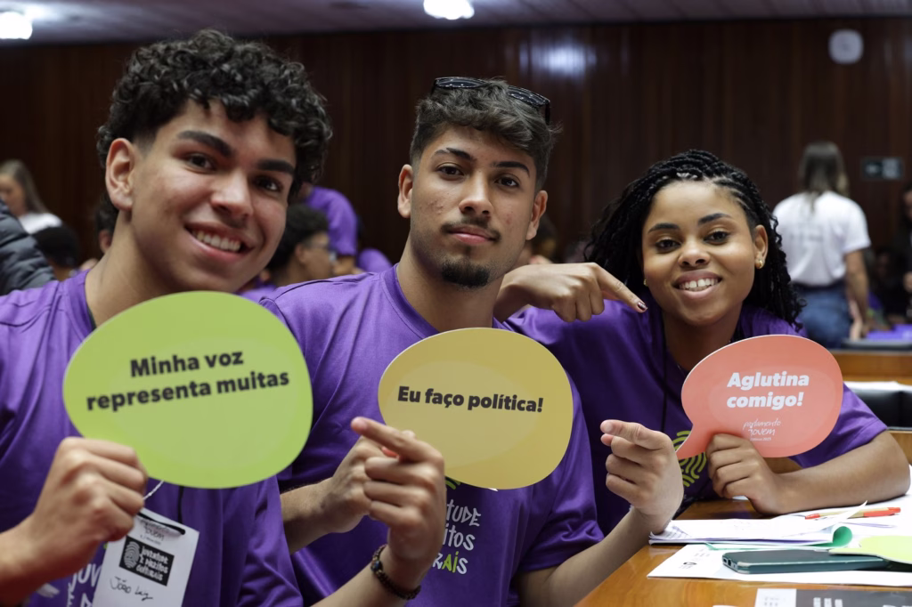  na imagem há três jovens sentados enfileirados no Plenário da Assembleia Legislativa de Minas Gerais. Dois desses jovens são homens que seguram placas contendo as frases “Minha voz representa muitas” e “Eu faço política!”, respectivamente. Há também uma mulher que sorri e aponta para a placa que segura em suas mãos, com a frase “Aglutina comigo!”. Os três usam a camiseta na cor roxa da edição de 2025 do projeto Parlamento Jovem.