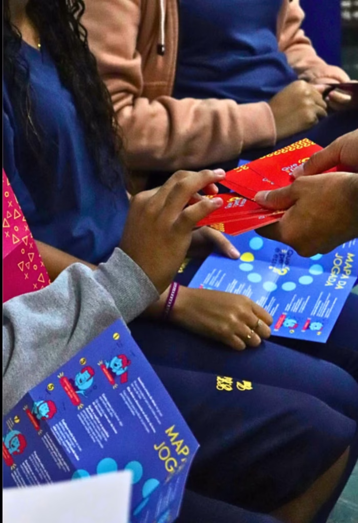 A foto mostra uma mão entregando materiais visivelmente educativos a várias pessoas sentadas, durante uma atividade educacional. Ela foca nas mãos de várias pessoas e em folhetos que estão sendo distribuídos. Vemos a mão de uma pessoa, segurando um folheto azul aberto, que diz "MAPA DA JOGADA" e textos abaixo.