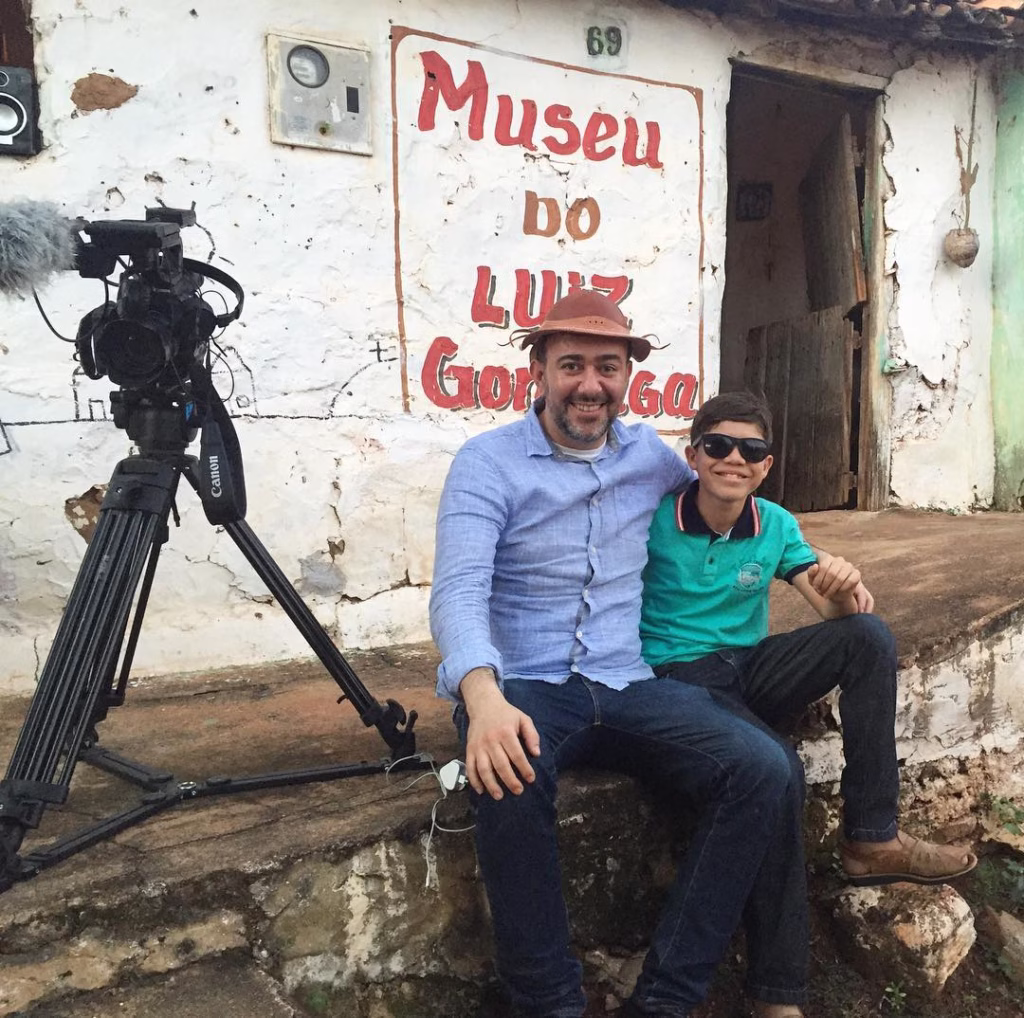 O jornalista Sérgio Utsch e um menino usando óculos escuros que criou um museu, eles estão sentados na frente de uma casa simples com a inscrição “Museu do Luiz Gonzaga” pintada na parede. Ao lado deles há uma câmera de vídeo montada em um tripé. A cena ocorre em um ambiente rural, com a fachada desgastada do prédio ao fundo.