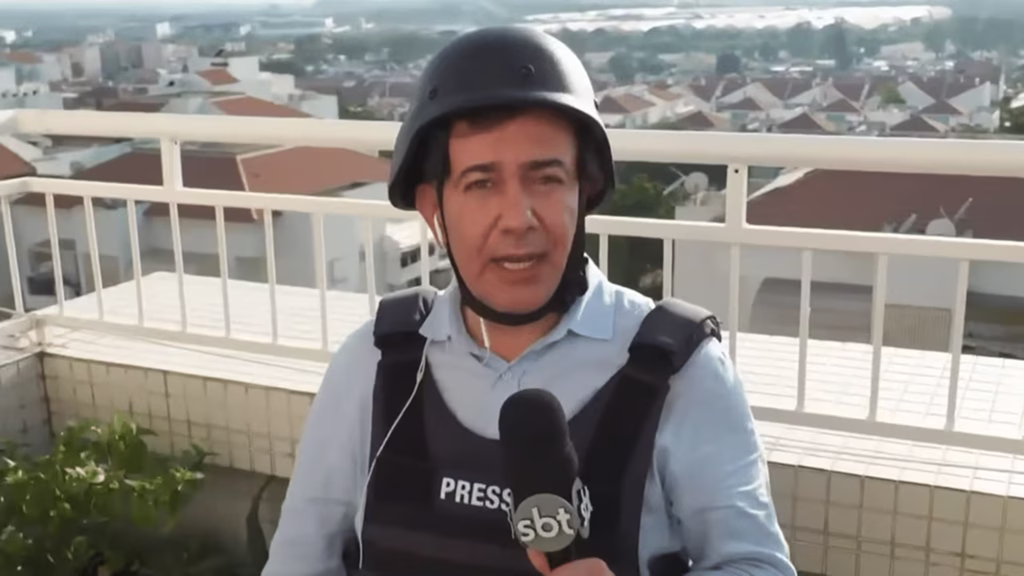 Na imagem, o jornalista Sérgio Utsch está em uma varanda ou cobertura de prédio, usando capacete preto e colete à prova de balas identificado com a palavra “Press”. Ele segura um microfone do SBT e usa fone de ouvido. Ao fundo, aparecem casas e edifícios sob a luz do dia.