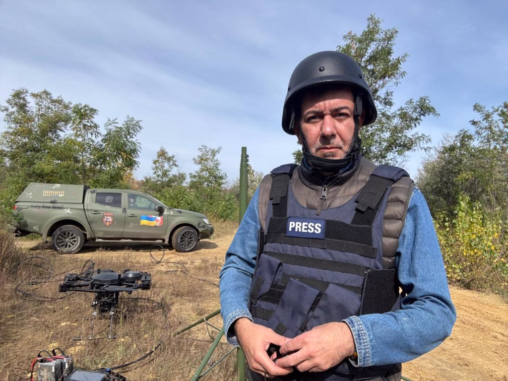 O jornalista Sérgio Utsch está em pé ao lado de um carro prata, usando óculos escuros, colete bege com bolsos e cachecol escuro. Ao fundo, há diversos caminhões estacionados em uma estrada cercada por montanhas rochosas, sob céu claro. Ele está voltado para a câmera, com uma das mãos apoiada na cintura.