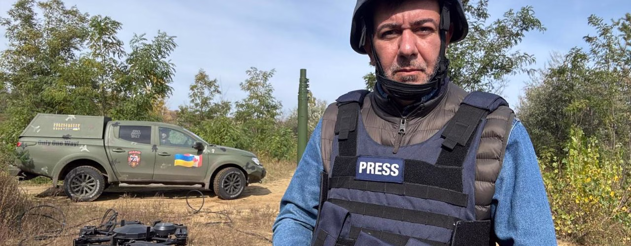 O jornalista Sérgio Utsch está em pé ao lado de um carro prata, usando óculos escuros, colete bege com bolsos e cachecol escuro. Ao fundo, há diversos caminhões estacionados em uma estrada cercada por montanhas rochosas, sob céu claro. Ele está voltado para a câmera, com uma das mãos apoiada na cintura.