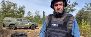 O jornalista Sérgio Utsch está em pé ao lado de um carro prata, usando óculos escuros, colete bege com bolsos e cachecol escuro. Ao fundo, há diversos caminhões estacionados em uma estrada cercada por montanhas rochosas, sob céu claro. Ele está voltado para a câmera, com uma das mãos apoiada na cintura.