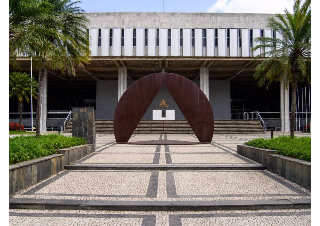Vista frontal da entrada da Assembleia Legislativa de Minas Gerais. Em primeiro plano, uma grande escultura metálica em formato semicircular envolve um triângulo vazado, posicionada sobre um piso de pedras claras com faixas escuras. Ao fundo, o edifício de arquitetura modernista apresenta escadarias, colunas e fachada de concreto com janelas verticais. Palmeiras e jardins ladeiam a entrada, sob céu parcialmente nublado.