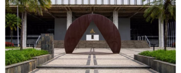 Vista frontal da entrada da Assembleia Legislativa de Minas Gerais. Em primeiro plano, uma grande escultura metálica em formato semicircular envolve um triângulo vazado, posicionada sobre um piso de pedras claras com faixas escuras. Ao fundo, o edifício de arquitetura modernista apresenta escadarias, colunas e fachada de concreto com janelas verticais. Palmeiras e jardins ladeiam a entrada, sob céu parcialmente nublado.
