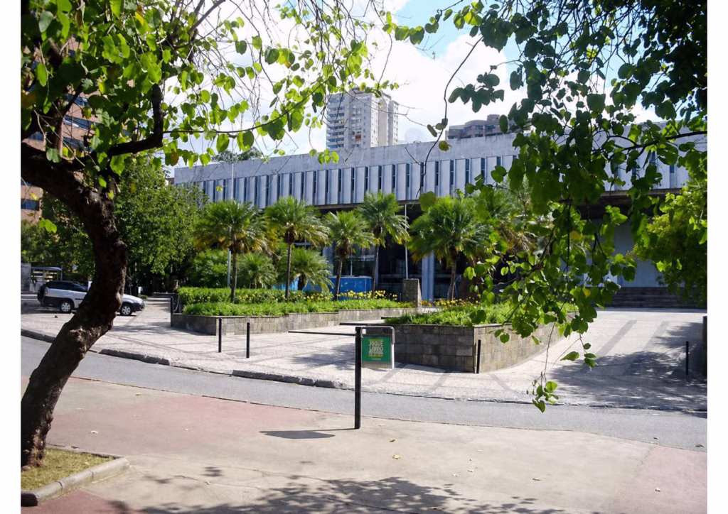 Fotografia ao ar livre mostrando a fachada de arquitetura moderna do prédio da ALMG (Assembleia Legislativa de Minas Gerais). A construção de concreto cinza e linhas verticais é precedida por uma praça com jardins, palmeiras e uma placa verde centralizada em primeiro plano. A cena é emoldurada por folhas de árvores nas bordas superior e esquerda.