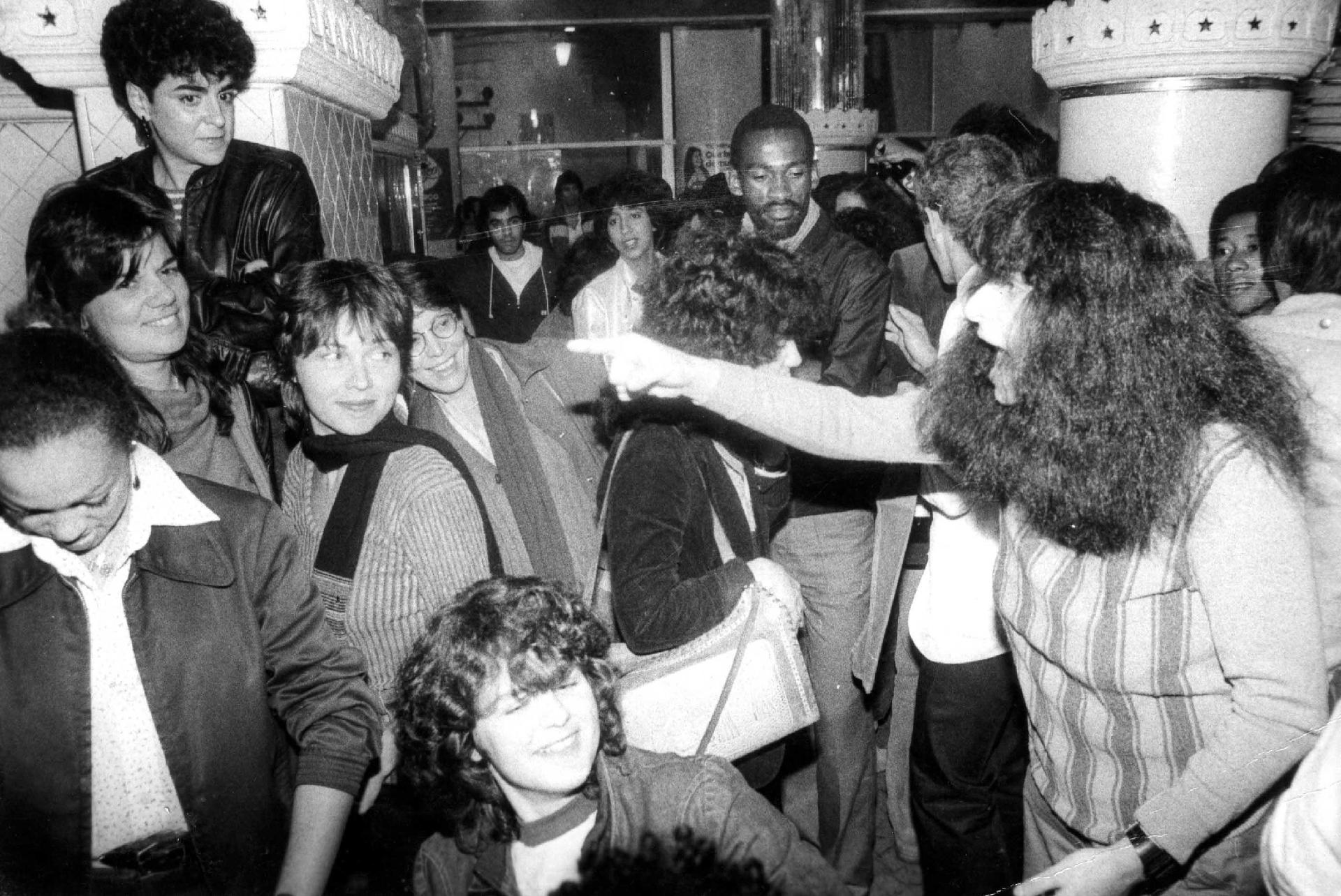 Momento do levante no Ferro's Bar. Na imagem em preto e branco, há cerca de 20 pessoas, em sua maioria mulheres, observando o discurso de Rosely Roth, à direita, que está fazendo um discurso caloroso enquanto gesticula.